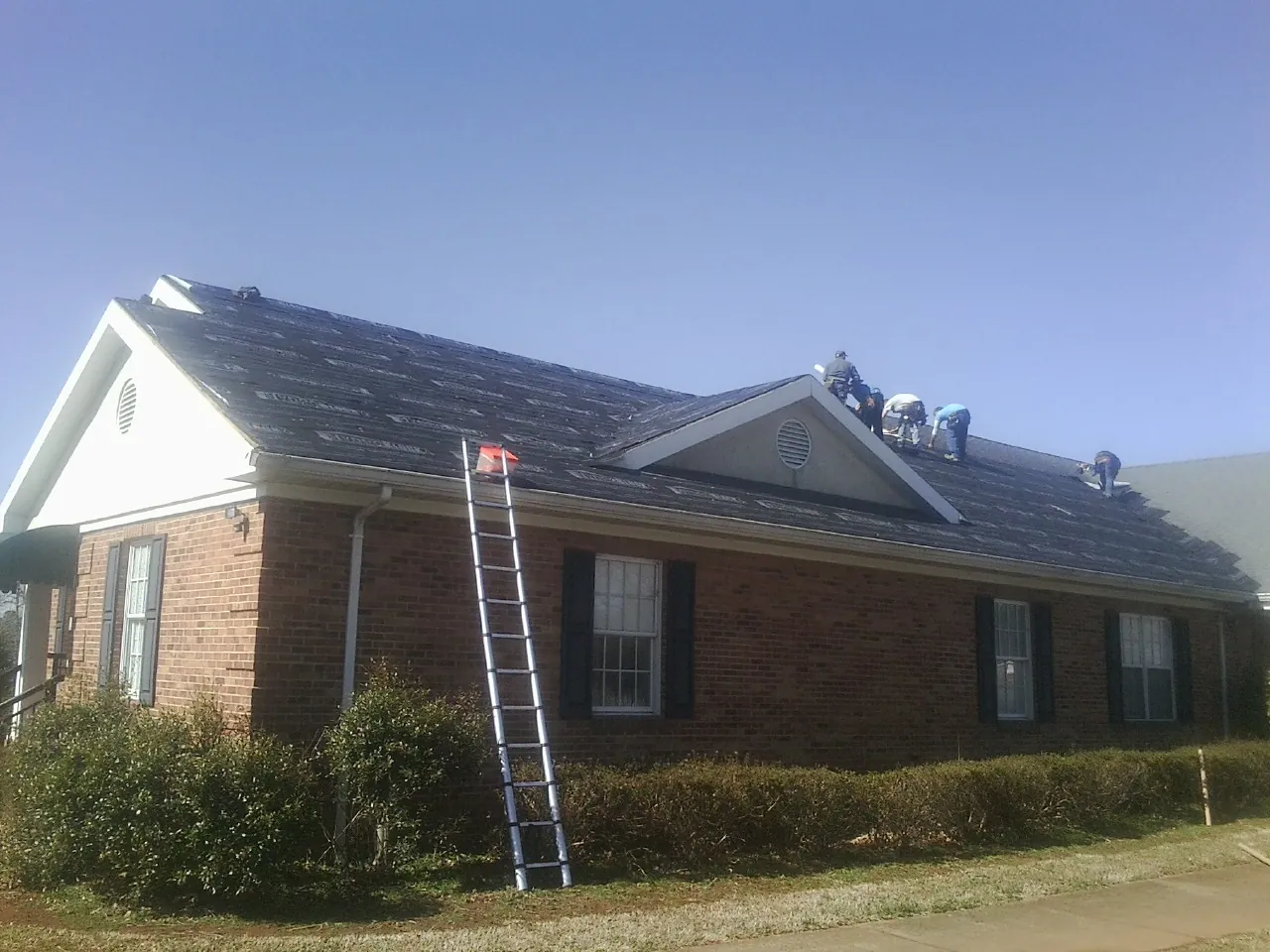 Crew tearing shingles off a brick ranch roof with a ladder set — Emergency Roof Repair work by our crew in Peru